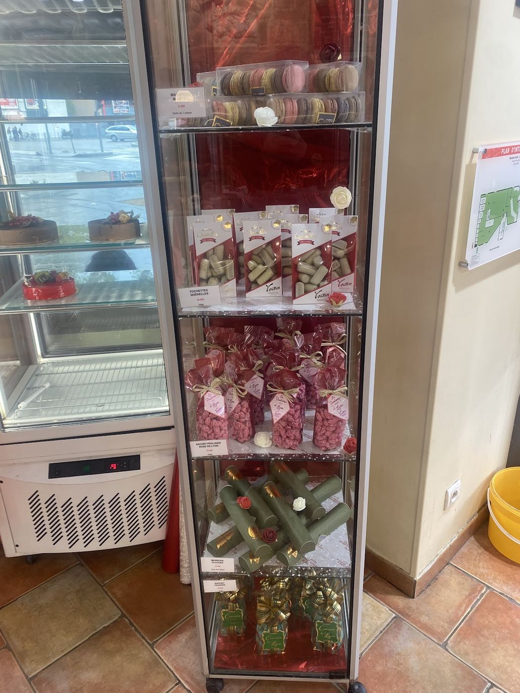 La vitrine à spécialités : macarons en boîte, pochettes de quenelles Voisin, sachets de pralines roses de Lyon nouées de raphia, marrons en étuis verts et coffrets dorés Coucou de Lyon, sur cinq étagères vitrées.