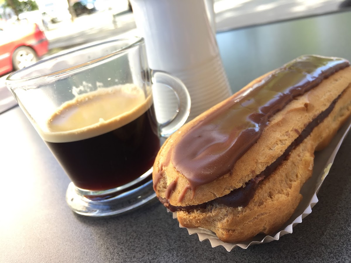 Un espresso servi en verre et un éclair au chocolat dans sa caissette, posés sur le comptoir devant la vitrine donnant sur l'avenue.