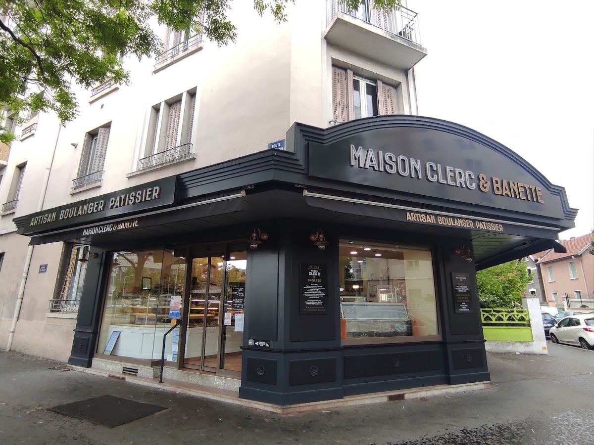 La façade à l'angle de l'avenue Paul Santy : marquise Art déco anthracite portant MAISON CLERC en lettres crème en relief et & BANETTE en lettres laiton, lampes en cuivre de part et d'autre de l'entrée vitrée.
